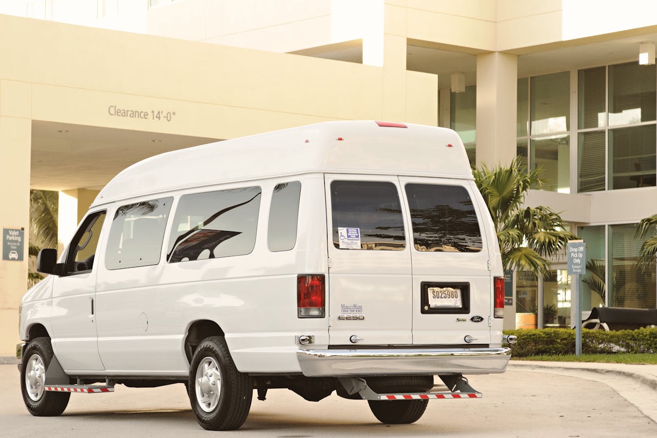 White passenger van parked in urban drop-off zone with palm trees and modern architecture.